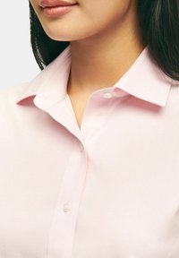 Chemise boutonnée rose clair avec un col structuré, présentant une texture douce et tissée, et une fermeture à un bouton au niveau du col.