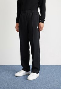 Svarta träningsbyxor med elastisk midja, sidofickor med dragkedja och vit Adidas-logotyp på vänster lår. Matchas med vita sneakers.