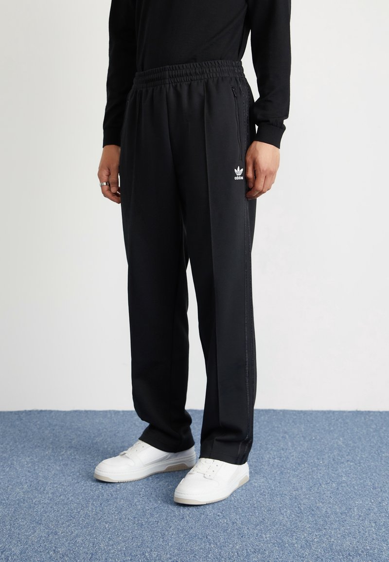 Svarta träningsbyxor med elastisk midja, sidofickor med dragkedja och vit Adidas-logotyp på vänster lår. Matchas med vita sneakers.