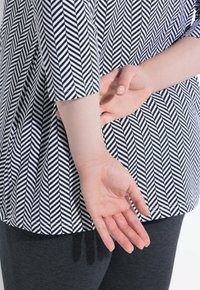 Zwart-witte top met chevronpatroon en 3/4 mouwen, gecombineerd met donkergrijze leggings. Zichtbare hand achter de rug geplaatst.