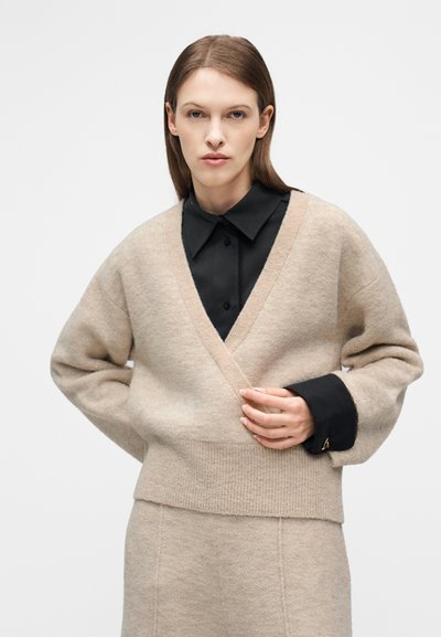 Γυναίκα που φοράει μπεζ πλεκτή ζακέτα και παντελόνι που ταιριάζουν, πάνω από μαύρο πουκάμισο με γιακά, κοιτάζοντας μπροστά και με το ένα χέρι να διορθώνει τη ζακέτα.