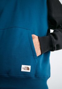 Mano nella tasca a canguro di una felpa blu e nera della North Face con il logo visibile sulla tasca.