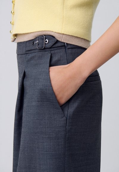 I pantaloni grigi sartoriali presentano un design elegante con una tasca laterale, accentuato da una cintura e una finitura testurizzata. Top a strati in maglina gialla e beige.