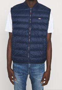 Hombre con chaleco acolchado azul marino sobre una camiseta blanca de manga corta y vaqueros azules, de pie con los brazos relajados a los lados.