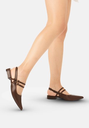 Benen met bruine puntige slingback platte schoenen met dubbele gespen, één voet opgetild halverwege de stap op een lichtgrijze achtergrond.