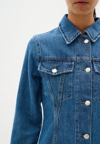 Veste en denim bleu avec un col pointu, des boutons métalliques et deux poches poitrine avec des détails de couture. Design et texture classiques.