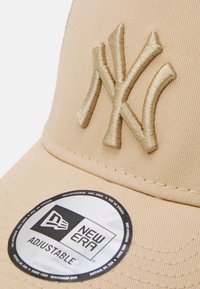 Nastavljiva bež kapa iz teksturiranega blaga, ki ima embosiran zlat NY logo in okroglo oznako New Era na robu.