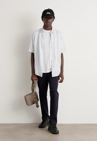 Kortermet, lyseblå rutete skjorte over en hvit t-skjorte, parret med marineblå bukser og svarte joggesko, holder en beige skulderbag.