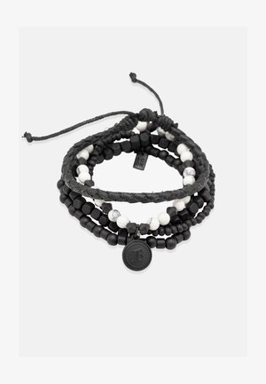 Flerlaget armbånd med sorte og hvide perler, flettet sort snor og en rund, præget charms. Inkluderer glatte og teksturerede overflader.