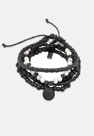 Mehrlagiges Armband mit schwarzen und weißen Perlen, geflochtenem schwarzen Band und einem runden, geprägten Charm. Enthält glatte und strukturierte Oberflächen.