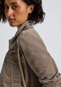 Kvinna med kort lockigt hår, iförd en taupe färgad corduroy jacka och bärande en beige axelväska, som tittar över sin vänstra axel.