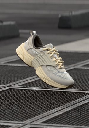 Baskets athlétiques beige clair et gris avec une semelle épaisse flottant au-dessus d'une grille métallique sur un trottoir urbain.