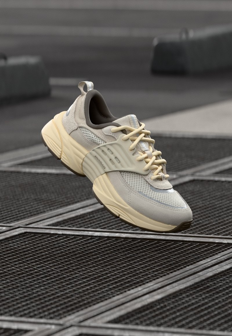 Baskets athlétiques beige clair et gris avec une semelle épaisse flottant au-dessus d'une grille métallique sur un trottoir urbain.