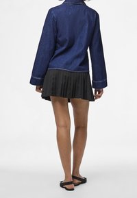 Jeansjacka i mörkblått med långa ärmar och sydda detaljer, kombinerad med en plisserad svart kjol och svarta flata sandaler.