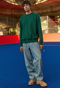 Groene sweatshirt, lichtblauwe baggy jeans en tan clogs. Model staat op een blauwe ondergrond, met een felrode achtergrond. Stedelijke omgeving, natuurlijk licht.