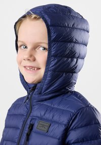 Tummansininen puhvijakku, jossa on vaakaraitoja, vetoketjukiinnitys ja kiristettävä huppu. Rintapielessä on logokoriste.
