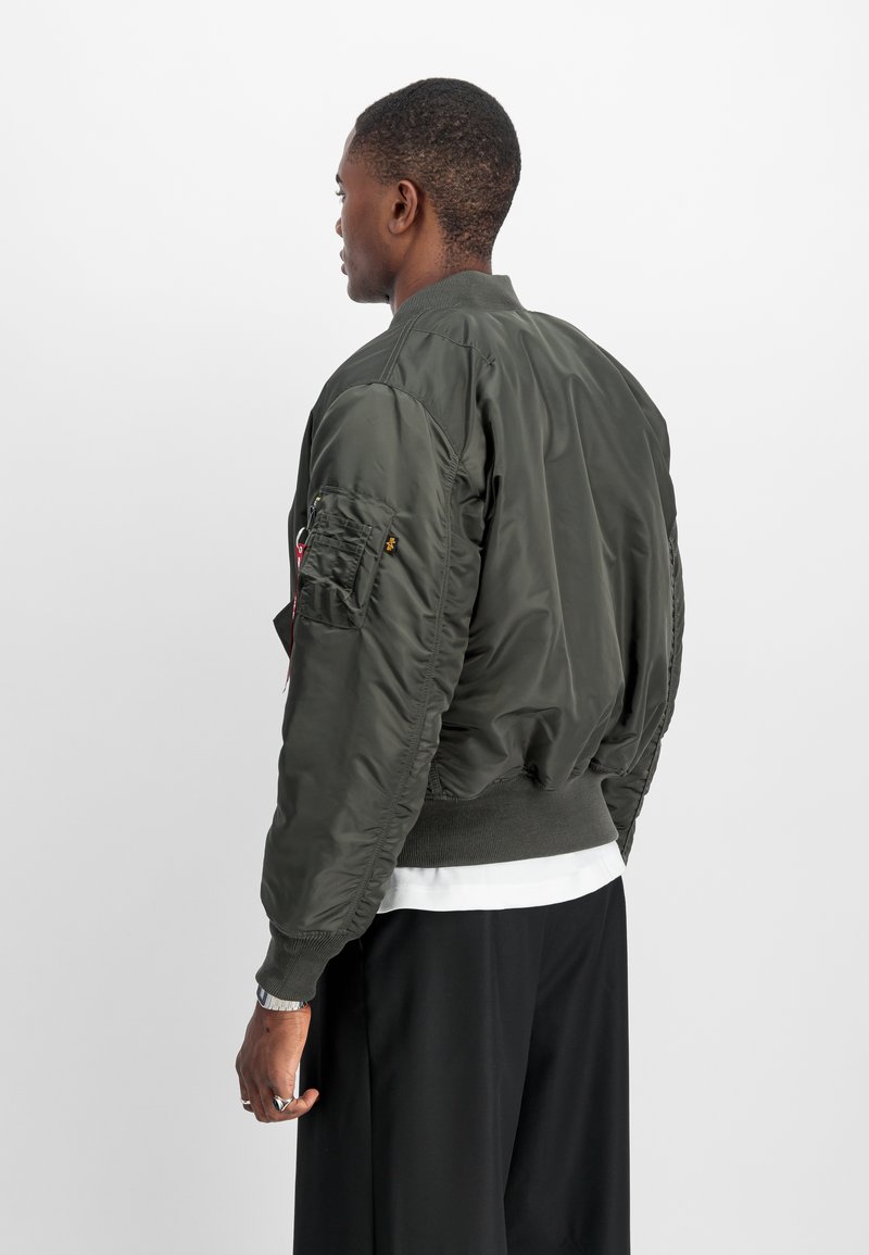 Chaqueta bomber de color verde oliva con textura acolchada, puños y cintura de canalé. Cuenta con un parche en el brazo izquierdo y un bolsillo con cremallera.