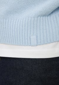Ourlet de pull tricoté bleu clair sur chemise blanche et jean en denim foncé, montrant la texture du tissu et une petite étiquette de vêtement.