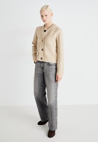 Beige cardigan med en teksturert finish, knappelukking og to lommeposer, kombinert med grå rette jeans og mørke støvler.