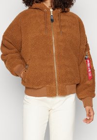 Hnědá mikina s fleece materiálem a zipem vpředu, oversized tvar, žebrované manžety a lem. Obsahuje detail červené nášivky a dvě boční kapsy.