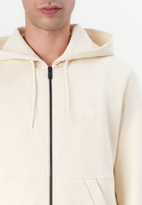 Lichtbeige zip-up hoodie met drawstrings, voorzien van een klein reliëflogo, voorzakken en geribbelde manchetten. Gladde textuur.