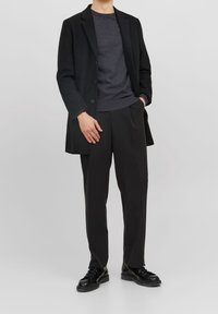 Svart kappa över en mörkgrå tröja, kombinerad med svarta byxor och svarta snörskor. Slät tyg, skräddarsydd passform och minimalistisk design.