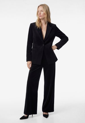 Femme blonde portant un costume en velours noir avec un blazer à un bouton et un pantalon à jambes larges, debout, une main sur la hanche.