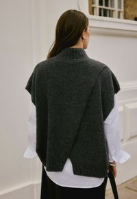 Woman with long brown hair wearing a dark gray sleeveless knit vest over a white long-sleeve shirt, holding a black strap, facing away.