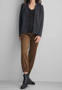 Veste en tissu gris mélangé avec encolure ronde et cinq boutons, associée à un pantalon jogger en satin marron et des bottines à lacets noires.