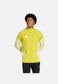 Gelbe Sportjacke mit weißen Akzenten, durchgehender Reißverschluss, Stehkragen und Adidas-Logo, aus glattem, leichtem Material.