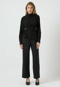 Haut noir à manches longues avec une conception croisée et un accent boucle ajustable, assorti à un pantalon noir à jambe droite et des escarpins à bout fermé.