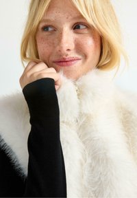 Valkoinen tekoturkisliivi, jossa on vetoketju, käytetään mustan pitkähihasen paidan päällä. Ominaisuuksiin kuuluu pehmeä rakenne ja korkea kaulus.