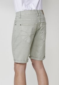 Vyras, apsirengęs šviesiai pilkiais trumpais šortais su apverstu kraštu ir baltu marškiniu, rodomas iš nugaros prieš vienodą foną.