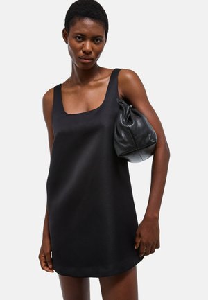 Femme aux cheveux courts portant une robe noire sans manches, tenant un sac en cuir noir sous son bras sur un fond uni.