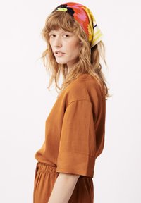 Een persoon draagt een roestkleurig shirt met korte mouwen en een elastische tailleband, aangevuld met een kleurrijke sjaal met een patroon.