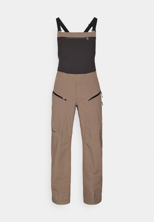 Braune und schwarze Outdoor-Latzhose mit verstellbaren Schulterträgern und mit Reißverschlüssen versehenen Taschen auf der Brust und den Oberschenkeln.