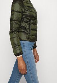Grön puffjacka med glänsande yta, med horisontell quiltning, ficka med dragkedja på sidan och ribbade ärmslut. Stylad med blåjeans.
