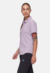 Camisa de manga corta a cuadros morados con diseño de botones. Presenta un dobladillo y un cuello redondeados, combinada con pantalones negros que muestran un logo.