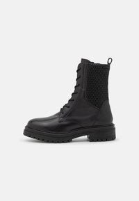 Bota de combate de cuero negro con suela gruesa, cordones en la parte delantera y panel trasero acolchado texturizado, presentada sobre un fondo blanco.