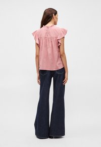 Ružičasta majica kratkih rukava s volanima i nabranim leđima, u paru s širokim tamnim trapericama. Glatka tekstura i suptilan uzorak.