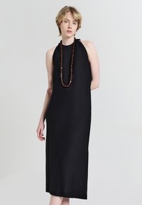 Schwarzes ärmelloses Kleid mit hohem Kragen, lockerer Passform, knielang und mit diagonaler Nahtdetails. Akzentuiert mit einer langen Perlenkette.