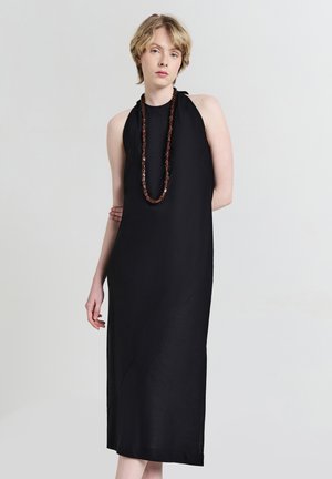 Schwarzes ärmelloses Kleid mit hohem Kragen, lockerer Passform, knielang und mit diagonaler Nahtdetails. Akzentuiert mit einer langen Perlenkette.
