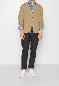 Beige stickad cardigan över en blårutig skjorta, mörka denimjeans och vita sneakers. Har knäppning fram och uppvikt ärmar.