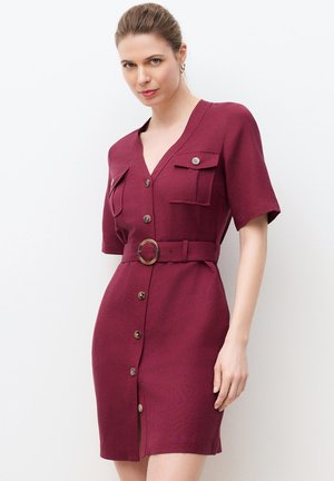 Kobieta w bordowej sukience z krótkim rękawem, zapinanej na guziki z paskiem i dwoma kieszeniami na piersi, stojąca na białym tle.
