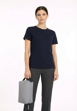 Vrouw met donker haar in een marineblauw T-shirt en grijze broek staat en houdt een grijze rechthoekige handtas vast aan de zwarte handvatten tegen een witte achtergrond.