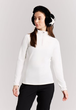 Ung kvinde iført sorte og hvide ørevarmere, hvid langærmet zip-up top og sorte bukser, stående mod en ensfarvet lys baggrund.