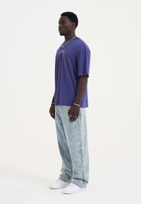 Paarse oversized T-shirt met korte mouwen, gecombineerd met lichtblauwe versleten jeans met een subtiel patroon, en witte sneakers.