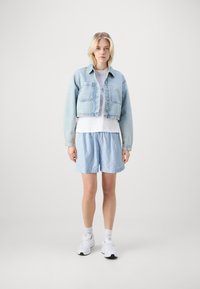 Veste en denim cropped bleu clair avec des boutons, associée à un t-shirt blanc et un short rayé bleu clair. Des baskets blanches complètent la tenue.