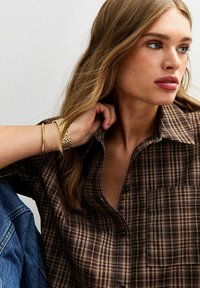 Brown plaid button-up shirt with a single pocket, collared design, and soft texture. Worn with a gold bracelet, and denim visible in the background.