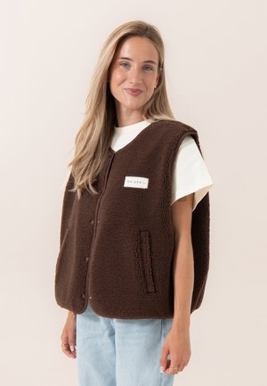 Vrouw met lang blond haar die een bruine mouwloze fleecevest draagt over een wit T-shirt met korte mouwen en lichtblauwe spijkerbroek, licht glimlachend.